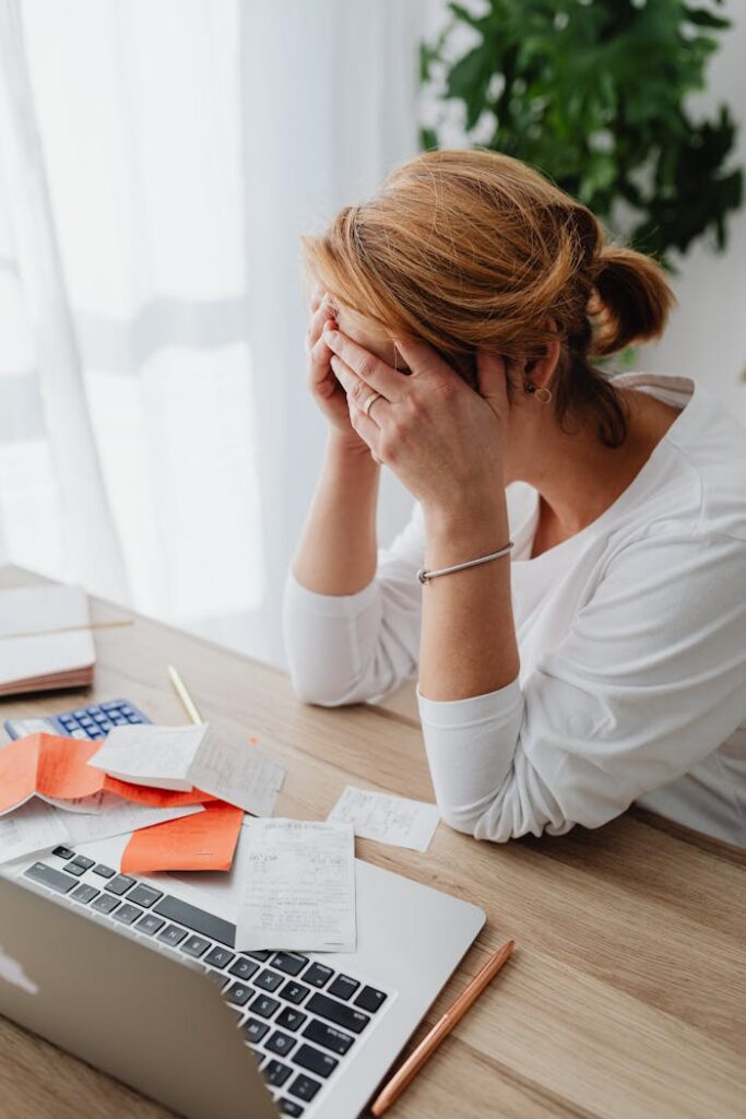 Overwhelmed woman sitting at desk with laptop and documents, showing stress and frustration.